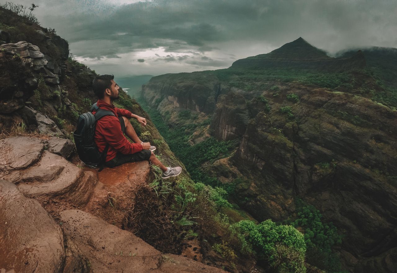 A man sits on a cliff edge, enjoying the scenic view of lush mountains and dramatic cloudy skies.