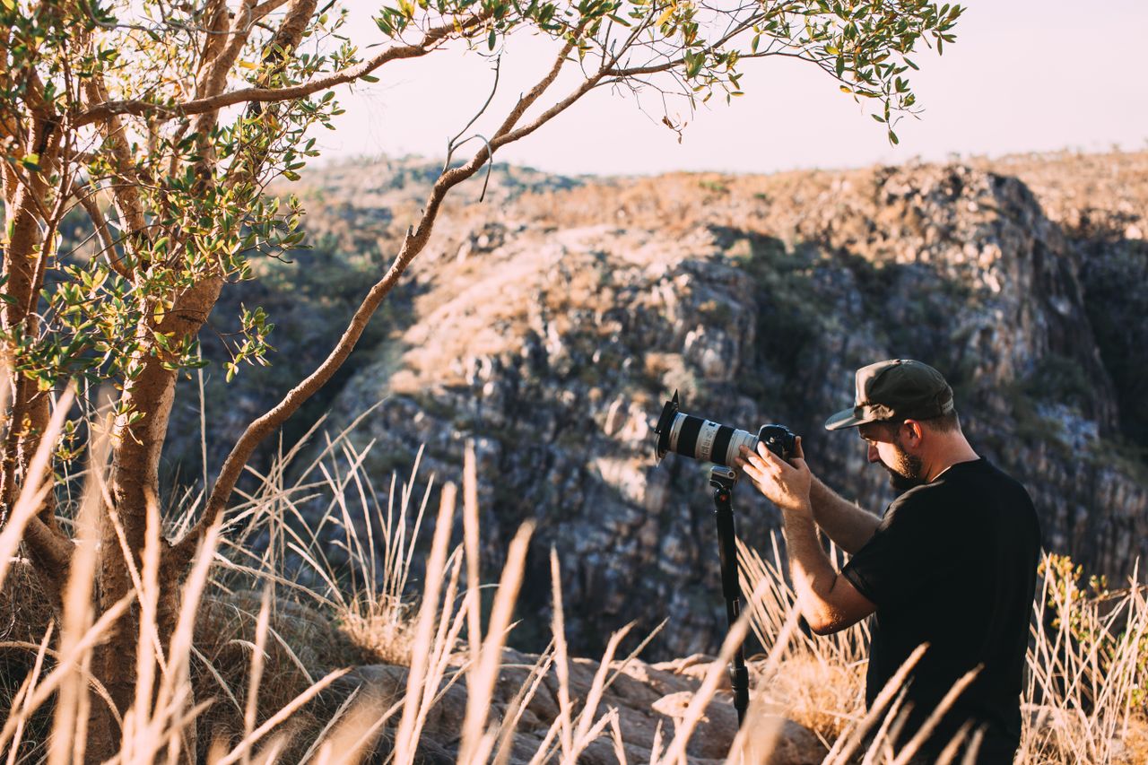 Side view of unrecognizable photographer in casual clothes and cap standing on grassy ground near green tree and taking photos of stone hills with photo camera in sunny summer day